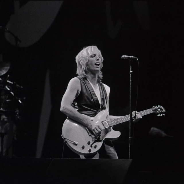 Monochrome photo of a fair-skinned adult male musician singing and playing an electric guitar on stage with a microphone.