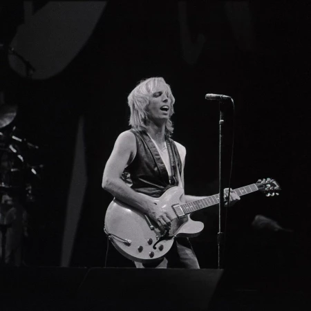 Monochrome photo of a fair-skinned adult male musician singing and playing an electric guitar on stage with a microphone.