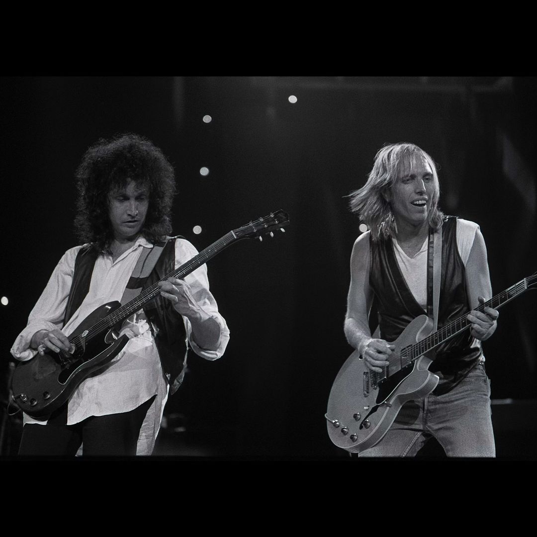 Two musicians perform with electric guitars on a dark stage with a spotlight backdrop, captured in a monochrome concert photograph.