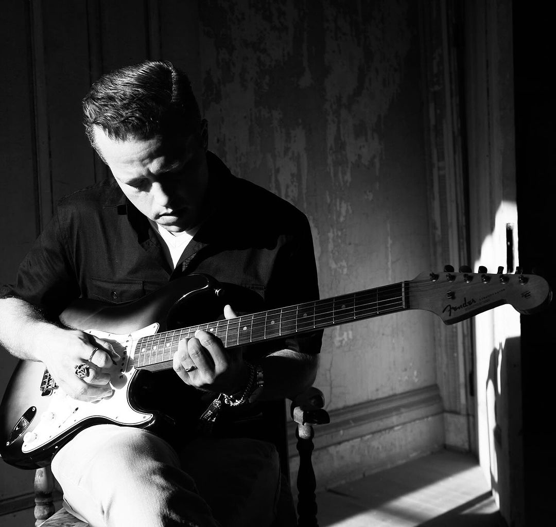 Black and white portrait of adult male playing electric guitar in a room with natural window light and cracked wall.