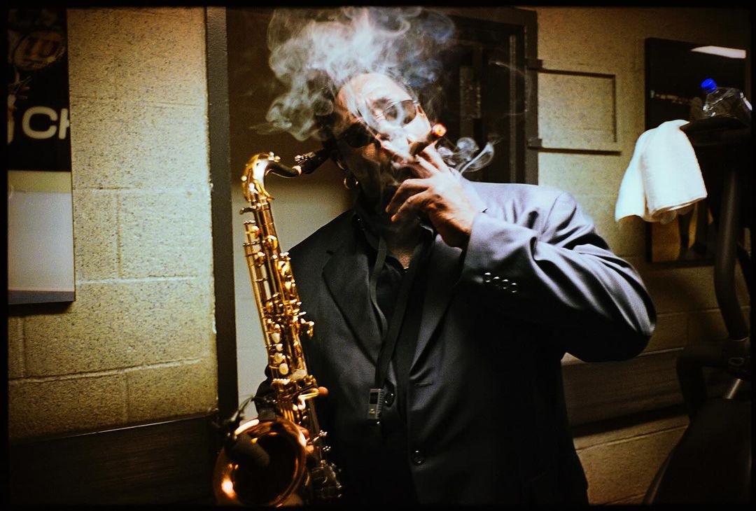 Musician smokes a cigar while holding a saxophone indoors in a dimly lit room with a brick wall and towel rack in the background.
