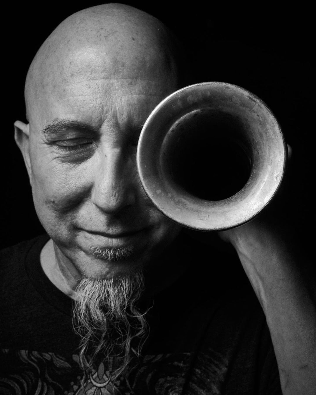 Black and white portrait of a bald adult male looking through the bell of a brass horn held up to his face in a dark studio.