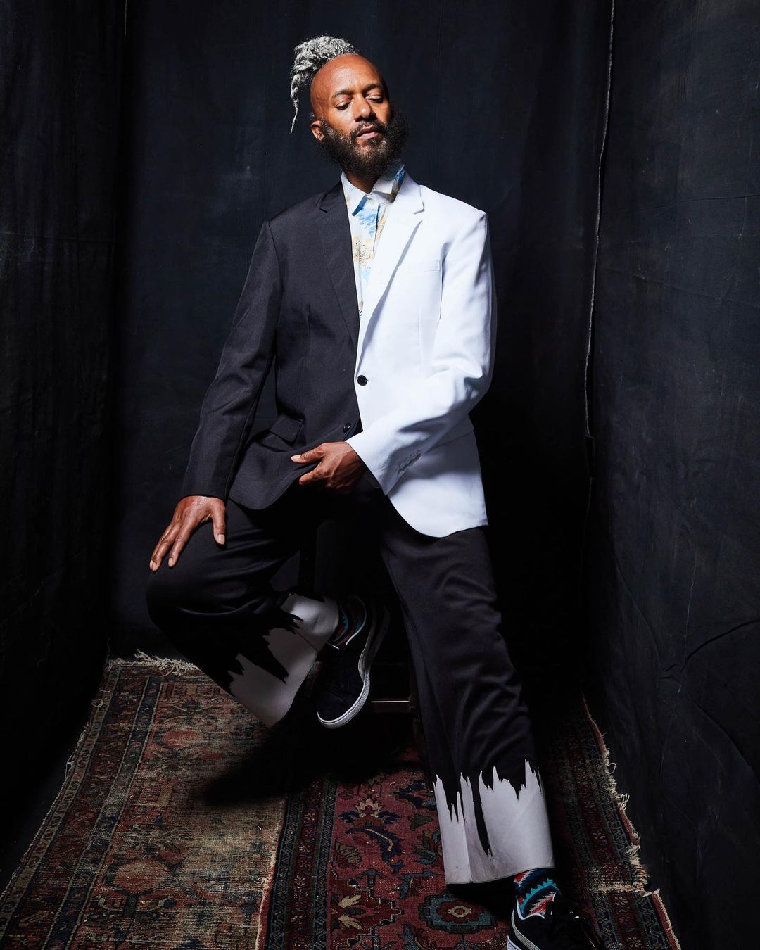 Stylish man in a unique half-black, half-white suit poses indoors with gray hair and beard, seated on a patterned oriental rug.