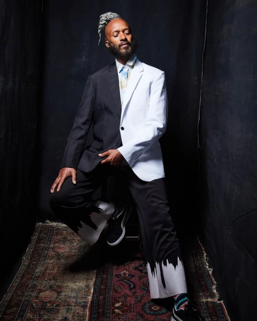 Stylish man in a unique half-black, half-white suit poses indoors with gray hair and beard, seated on a patterned oriental rug.