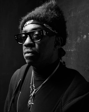 Portrait of a black man wearing dark glasses, a headband, and layered necklaces against a dark background by Danny Clinch.