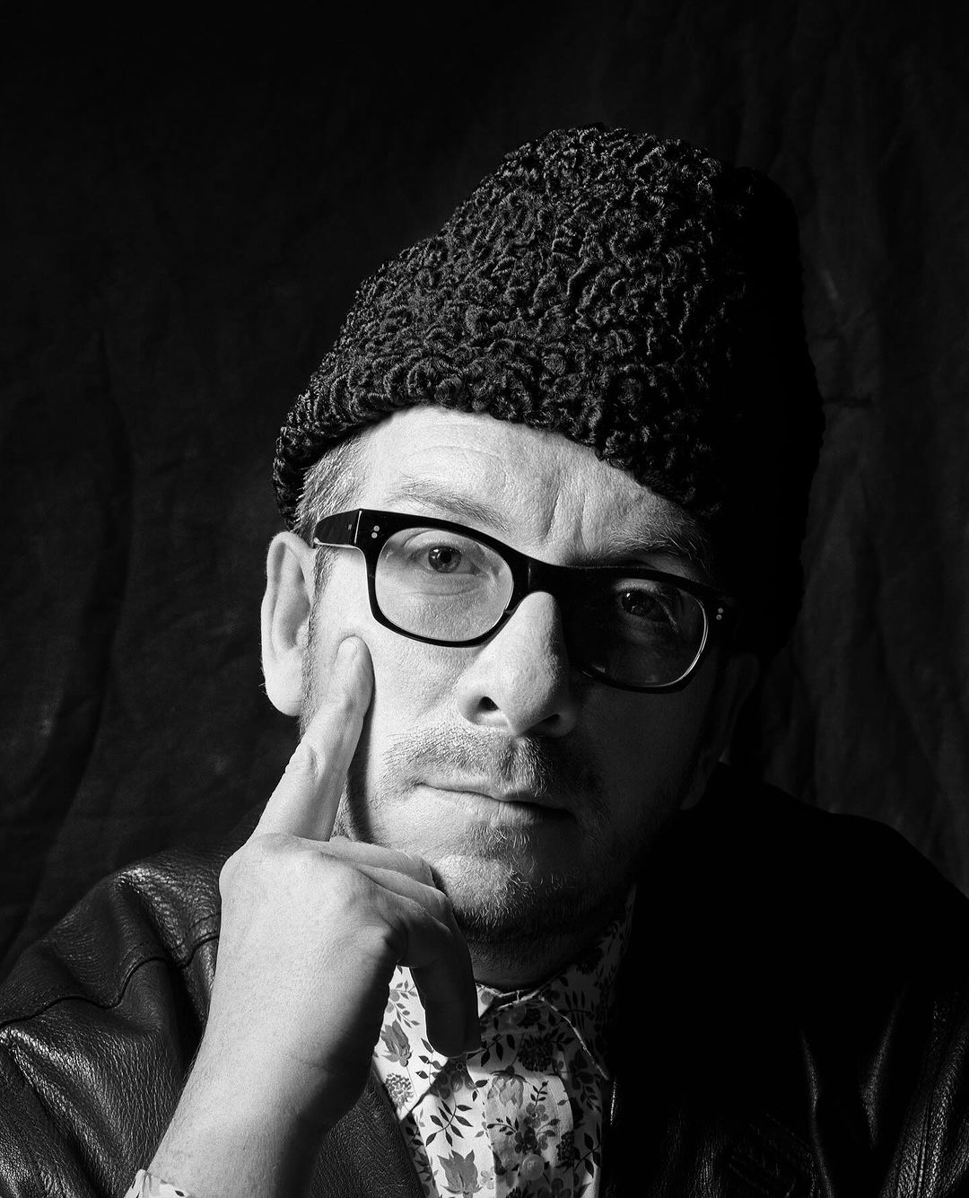 Black and white portrait of a man with glasses wearing a dark colored hat, leather jacket and floral shirt, with his hand on his face.