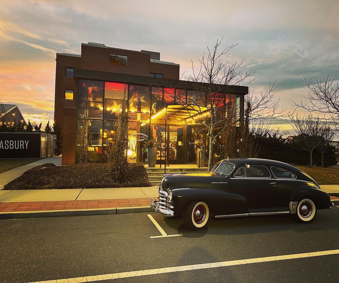 A vintage black car is parked in front of a modern brick building with large windows at sunset, creating a sense of classic elegance.