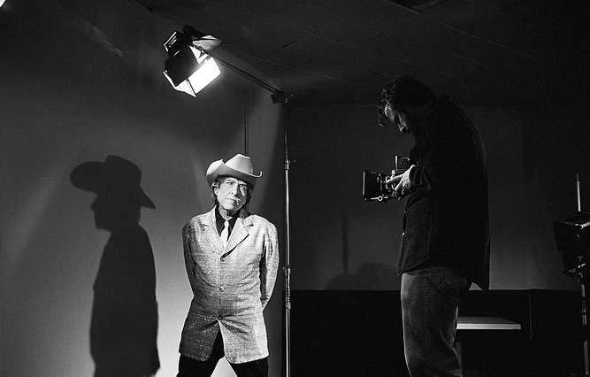 Black-and-white photo studio portrait of a mature adult in a cowboy hat and jacket, posing as a photographer aims a camera.