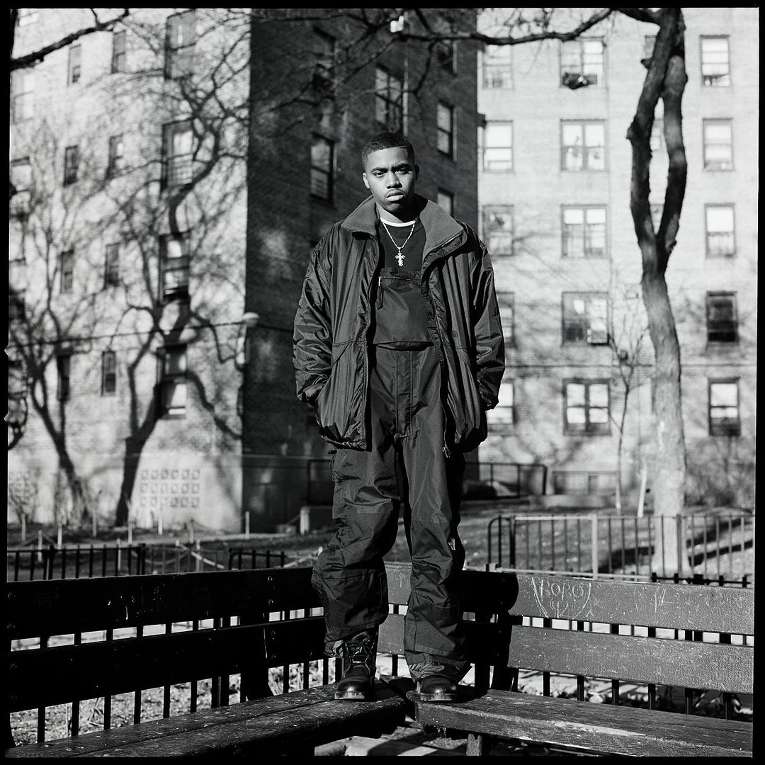 Black and white portrait of adult male standing on bench outdoors in jacket and boots, with apartment buildings and trees in background.