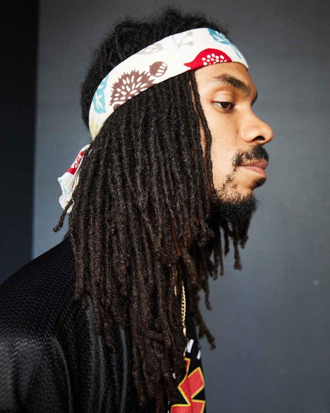 Portrait of adult male with dreadlocks, wearing a patterned headband indoors against a gray background, looking off to the side.