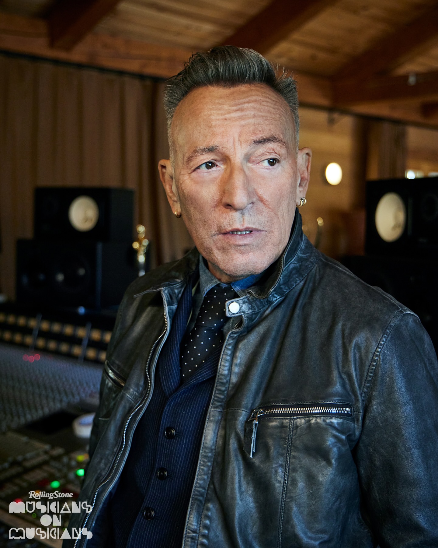 Portrait of Bruce Springsteen wearing a leather jacket, vest, and tie, indoors in a music studio with recording equipment visible.