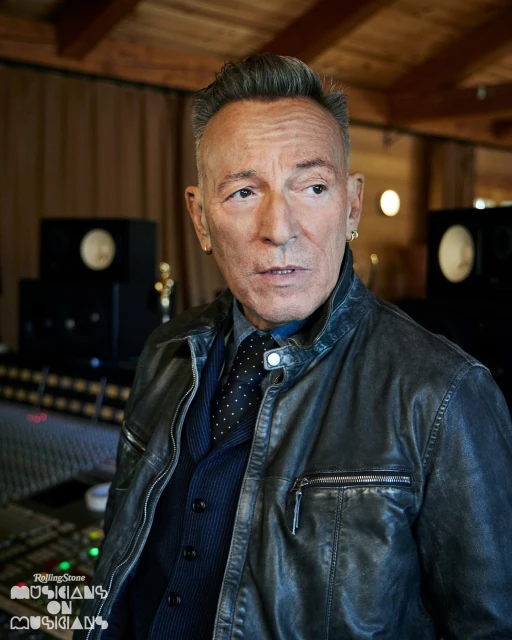 Portrait of Bruce Springsteen wearing a leather jacket, vest, and tie, indoors in a music studio with recording equipment visible.