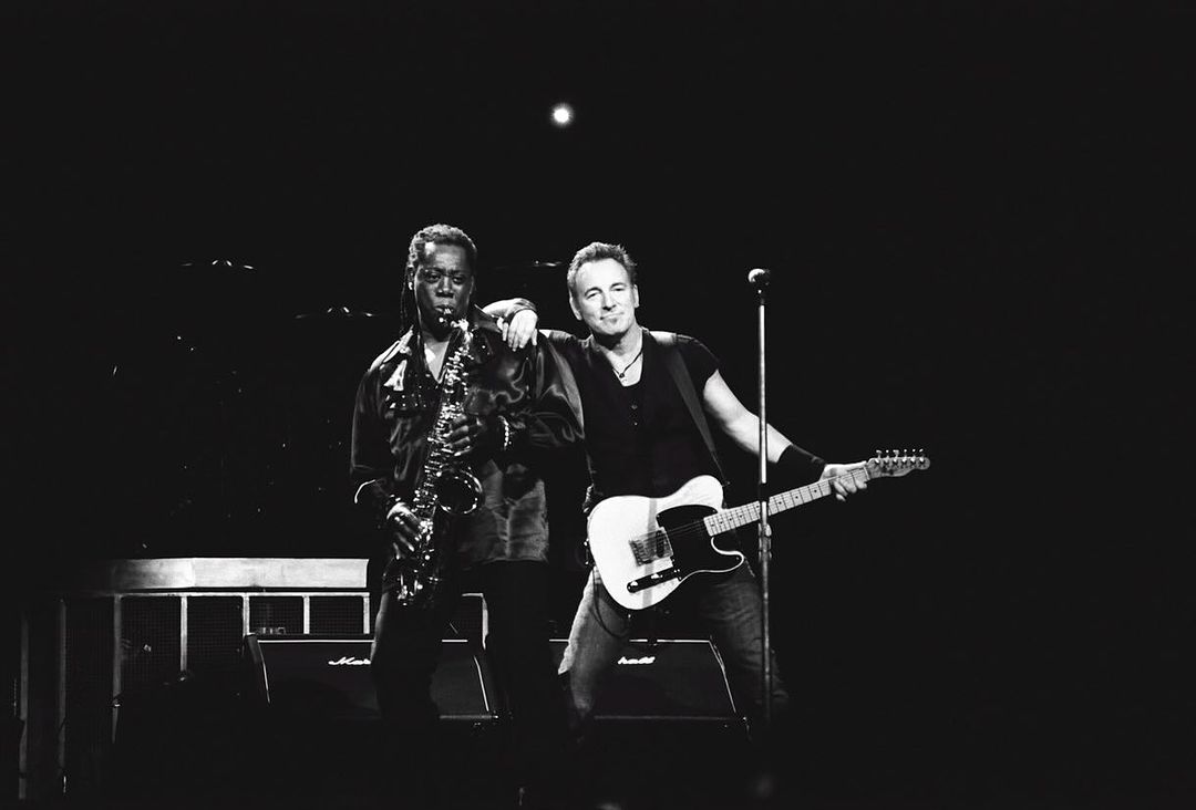 Black and white photo of Bruce Springsteen with a guitarist alongside Clarence Clemons playing a saxophone during live music performance.