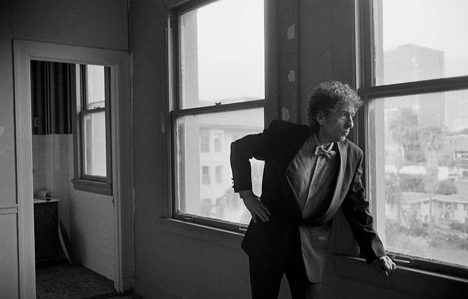 Black and white portrait of a man in a suit, standing indoors looking out a window, with cityscape visible in the background.
