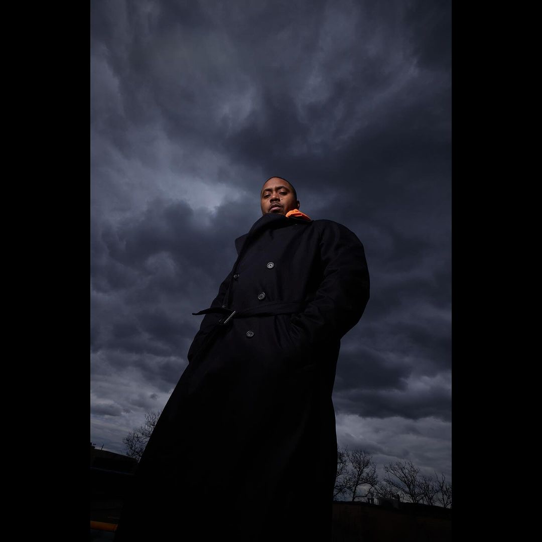 Man in a black coat standing outdoors against a cloudy sky, shot from a low angle, creating a dramatic portrait with a sense of depth.