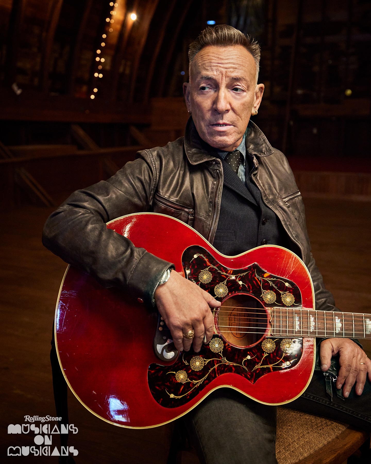 Portrait of Bruce Springsteen holding a red acoustic guitar, wearing a leather jacket, in a dimly lit room.