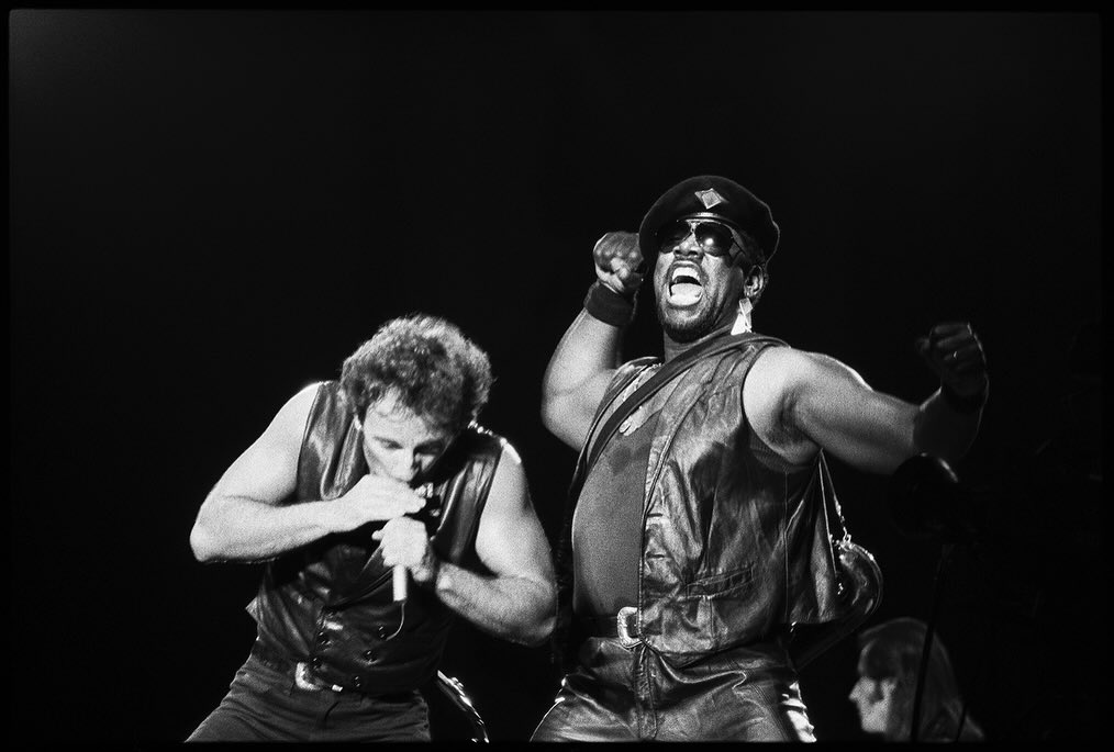 Two male musicians perform on stage in black vests and leather pants, singing into microphones with enthusiasm in a concert setting.