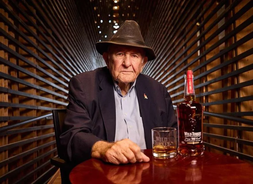 Elderly man in a hat sits at a table with a glass of whiskey and a bottle of Wild Turkey bourbon, posing indoors with wooden planks.