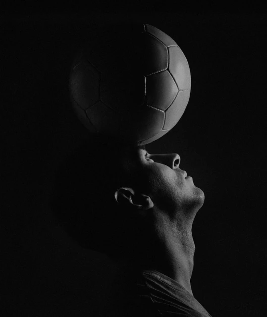 A man balances a soccer ball on his head in a dramatic black and white studio shot with strong lighting and a minimalist aesthetic.