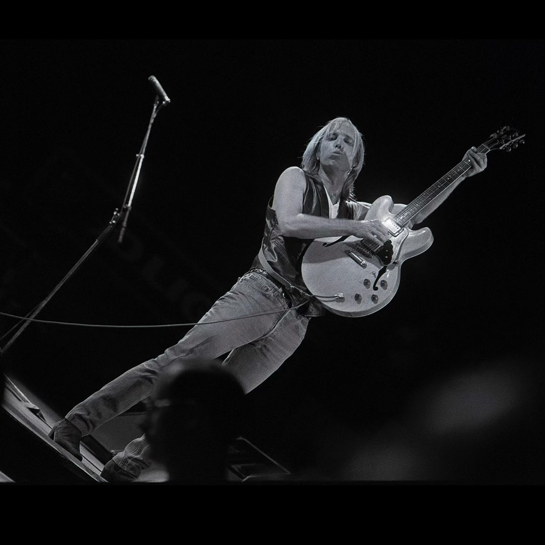 Monochrome concert photo of a musician playing a hollow-body electric guitar, singing into a microphone on a dark stage.