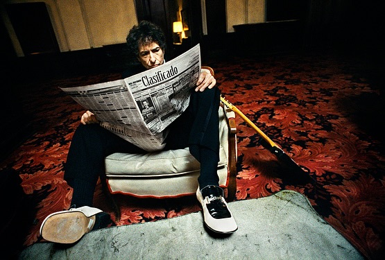 Man in chair reads newspaper with guitar in dimly lit room with patterned rug and dark walls, wearing black pants and white shoes.