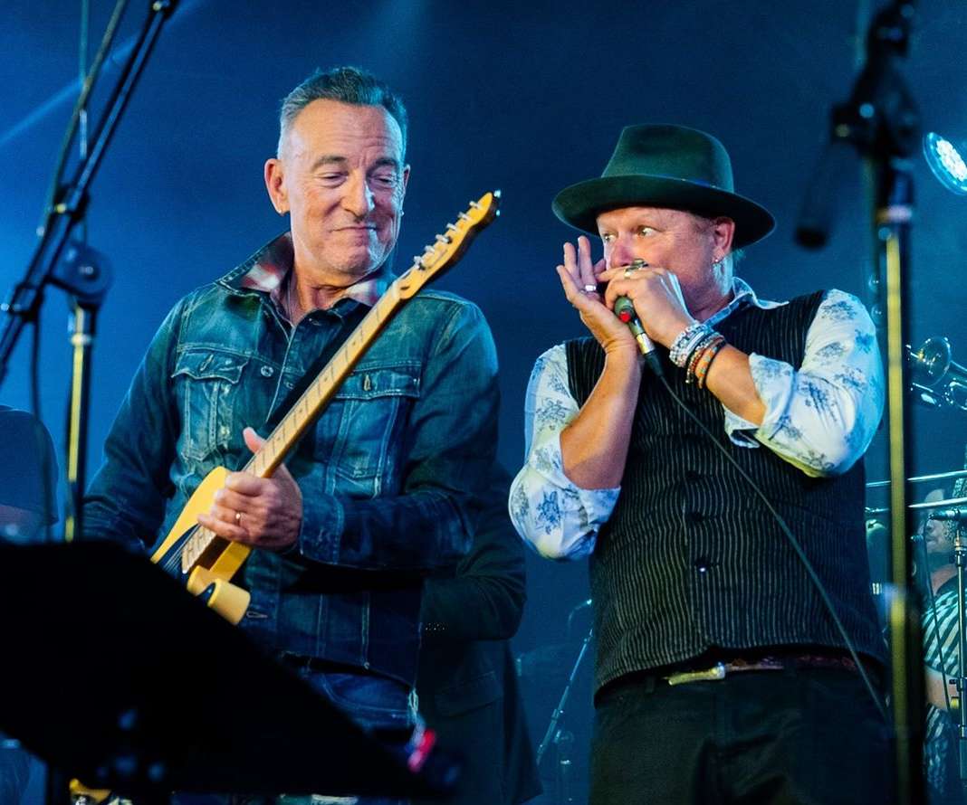 Two male musicians performing on stage at a concert; one playing guitar and singing, the other playing harmonica with band in background.