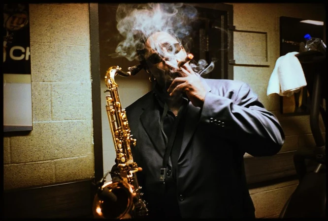 A man in a dark suit smoking a cigar while holding a golden saxophone in a backstage room.