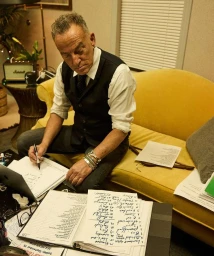 Bruce Springsteen seated on a yellow sofa writing on papers at a table, photographed by Danny Clinch in New York.