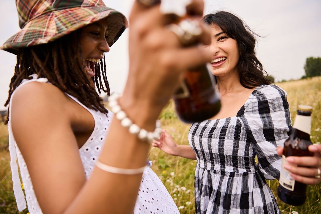 Two women laugh in a field, holding beer bottles in an outdoor setting. They wear summer dresses and a bucket hat, enjoying the moment.