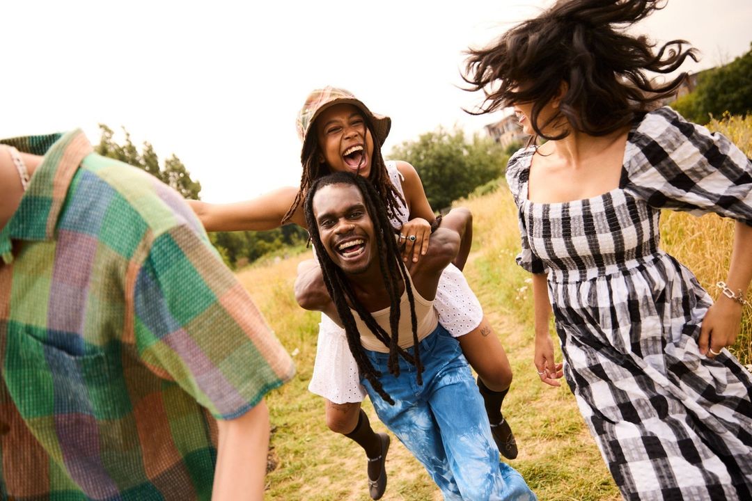 Group of young adults outdoors, smiling and laughing. One man carries a woman on his back. Friends wearing casual summer clothing.
