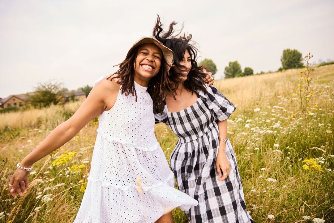 Two adult females embrace in a sunlit field with wildflowers. One wears a white dress and straw hat, while the other sports a checkered dress.