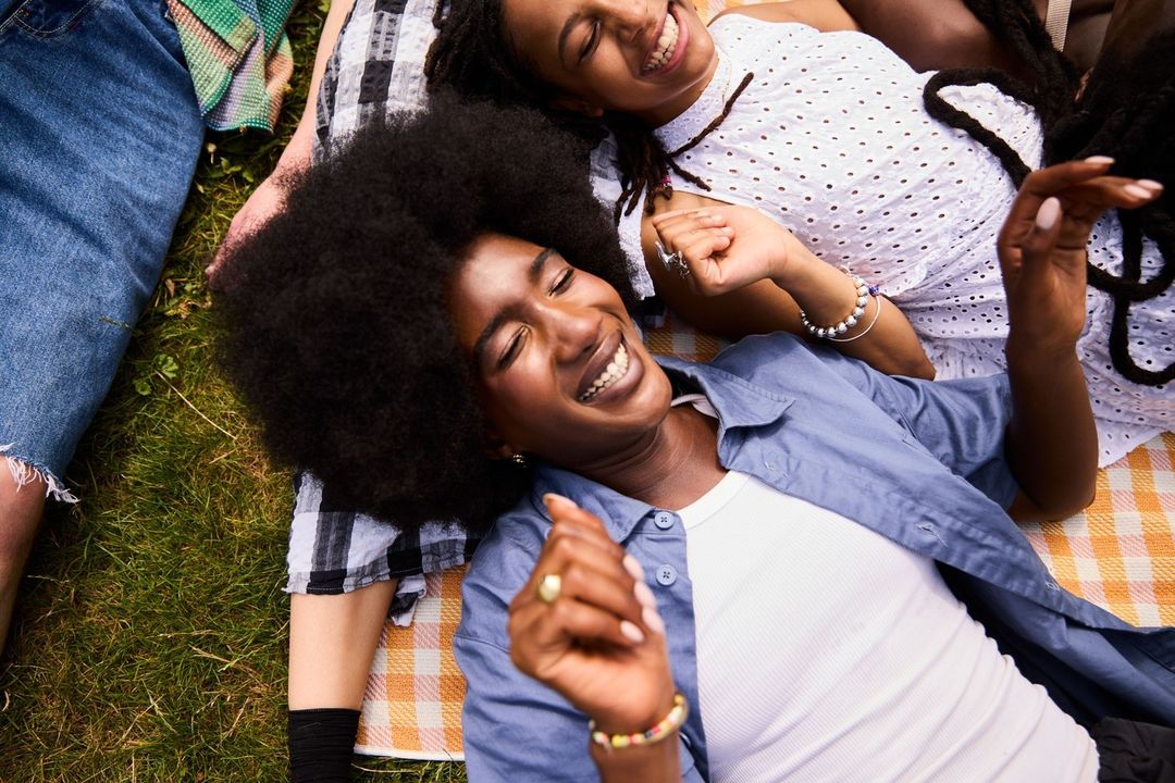 Two Black women smiling while lying on a picnic blanket on green grass. One woman wears a blue shirt. Summer lifestyle aesthetic.