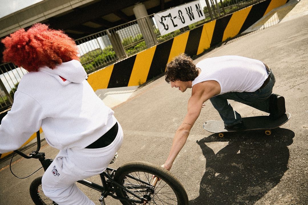 Two people, one on a skateboard and one on a bicycle, are the primary focus in an outdoor, urban setting with visible banner and shadows.