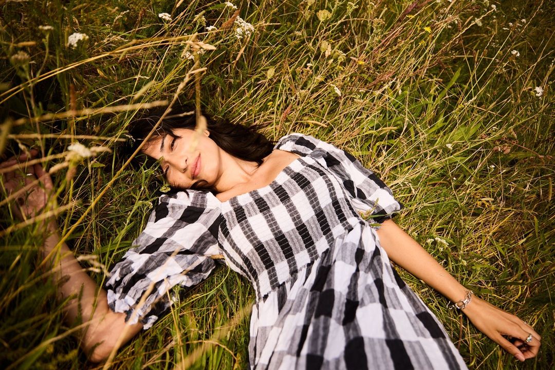 Brunette woman in a gingham dress relaxes in tall grass with white wildflowers, captured in a high-angle shot on a sunny day.