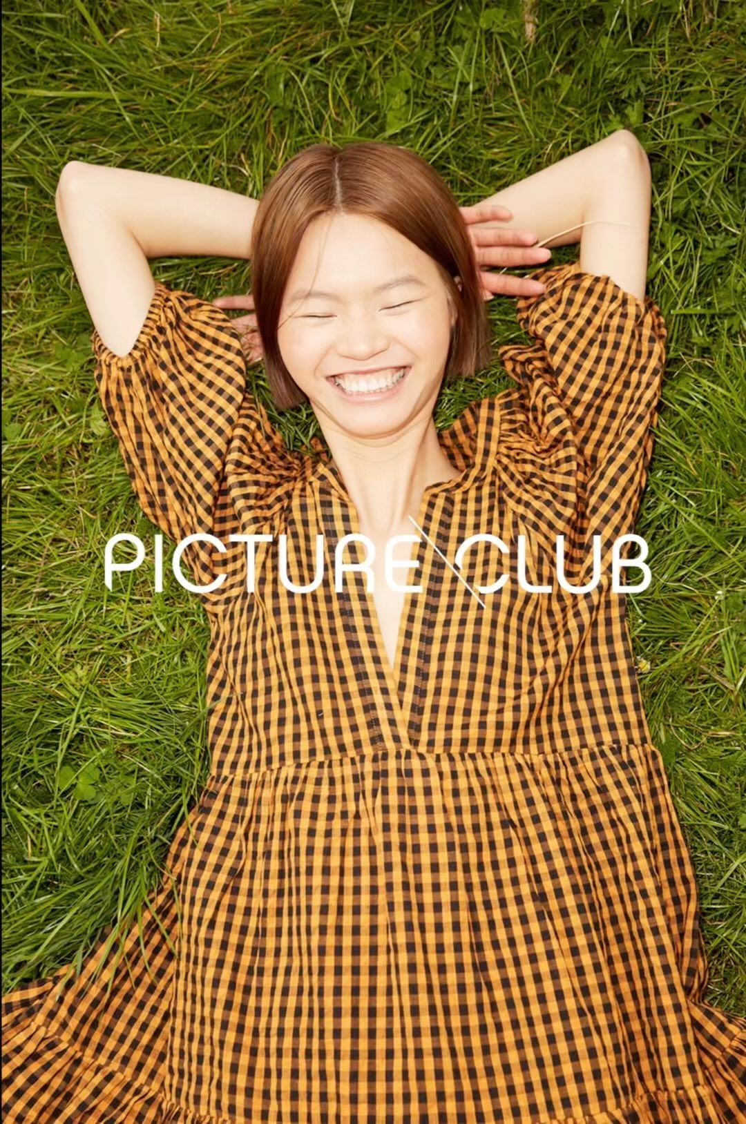 Young woman with short brown hair smiles, lying on her back in green grass wearing a plaid dress, looking directly at the camera.