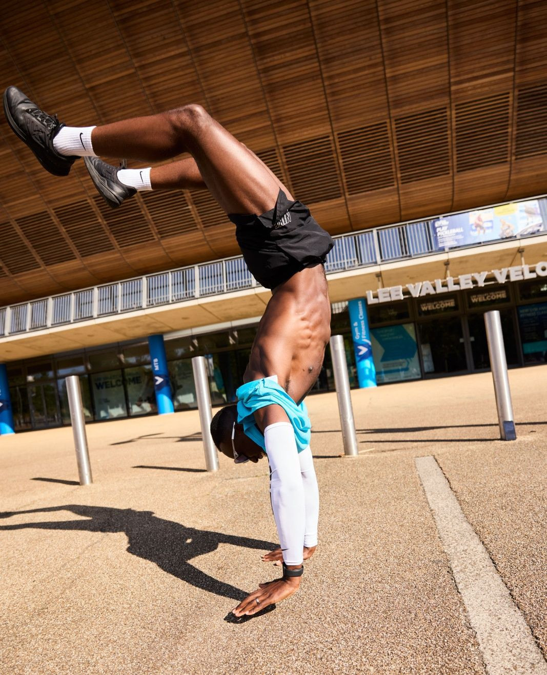 Athletic man doing a handstand outdoors in front of a building with brown wood paneling and silver columns on a sunny day.
