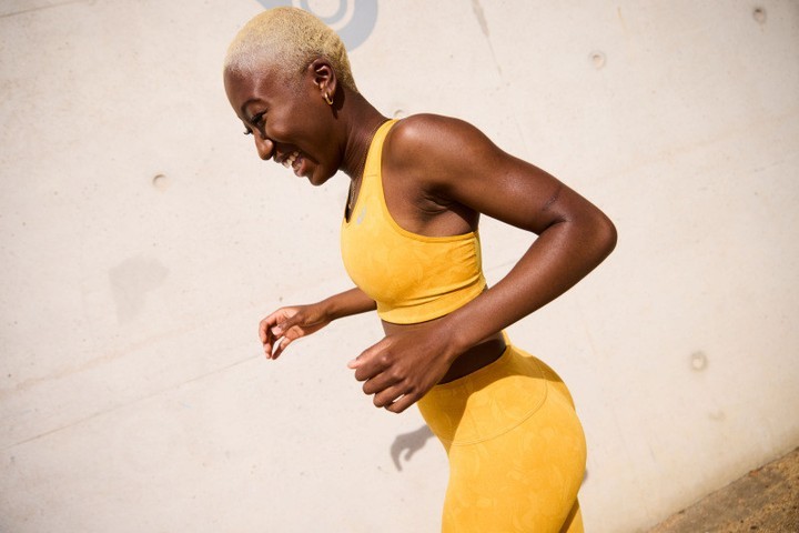 A smiling Black woman in a yellow sports bra and leggings poses against a concrete wall, showcasing athletic apparel in an active setting.