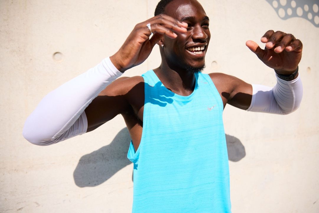 Smiling athletic man stands outdoors wearing a tank top and arm sleeves against a light-colored wall, enjoying the sunshine.