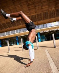 A shirtless black athlete performing a handstand outside a modern wooden building in London.
