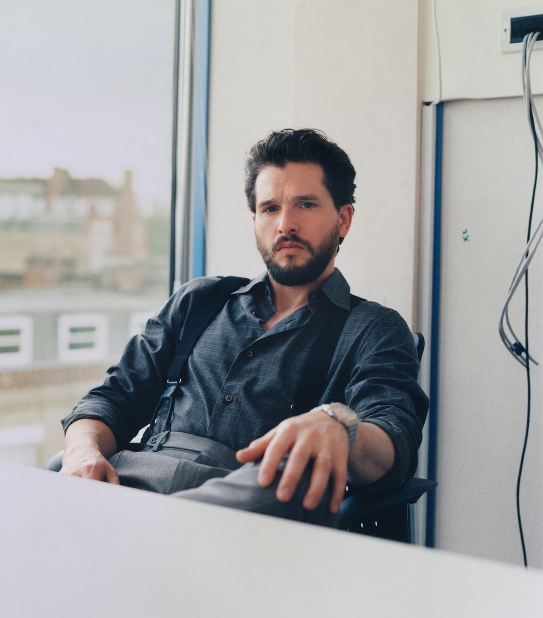Portrait of a dark-haired, bearded man in a button-down shirt and suspenders sitting at a desk in an office, gazing intently at the camera.