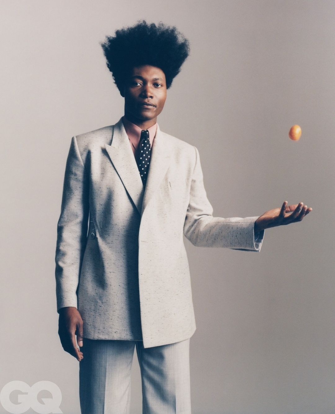 A stylish Black man in a suit poses in a studio, tossing an orange in the air, showcasing an elegant and fashionable look against a gray backdrop.