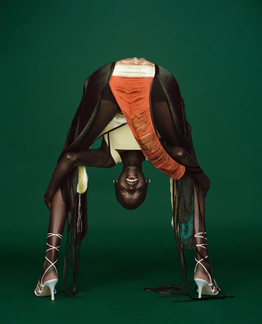 Adult female model bending over backwards in a studio. She is wearing a yellow top, white heels, and fringed clothing in a studio.
