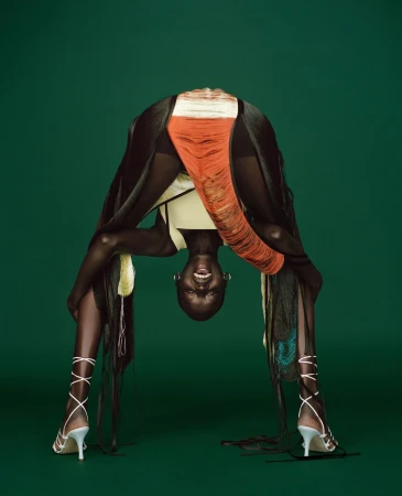 Adult female model bending over backwards in a studio. She is wearing a yellow top, white heels, and fringed clothing in a studio.