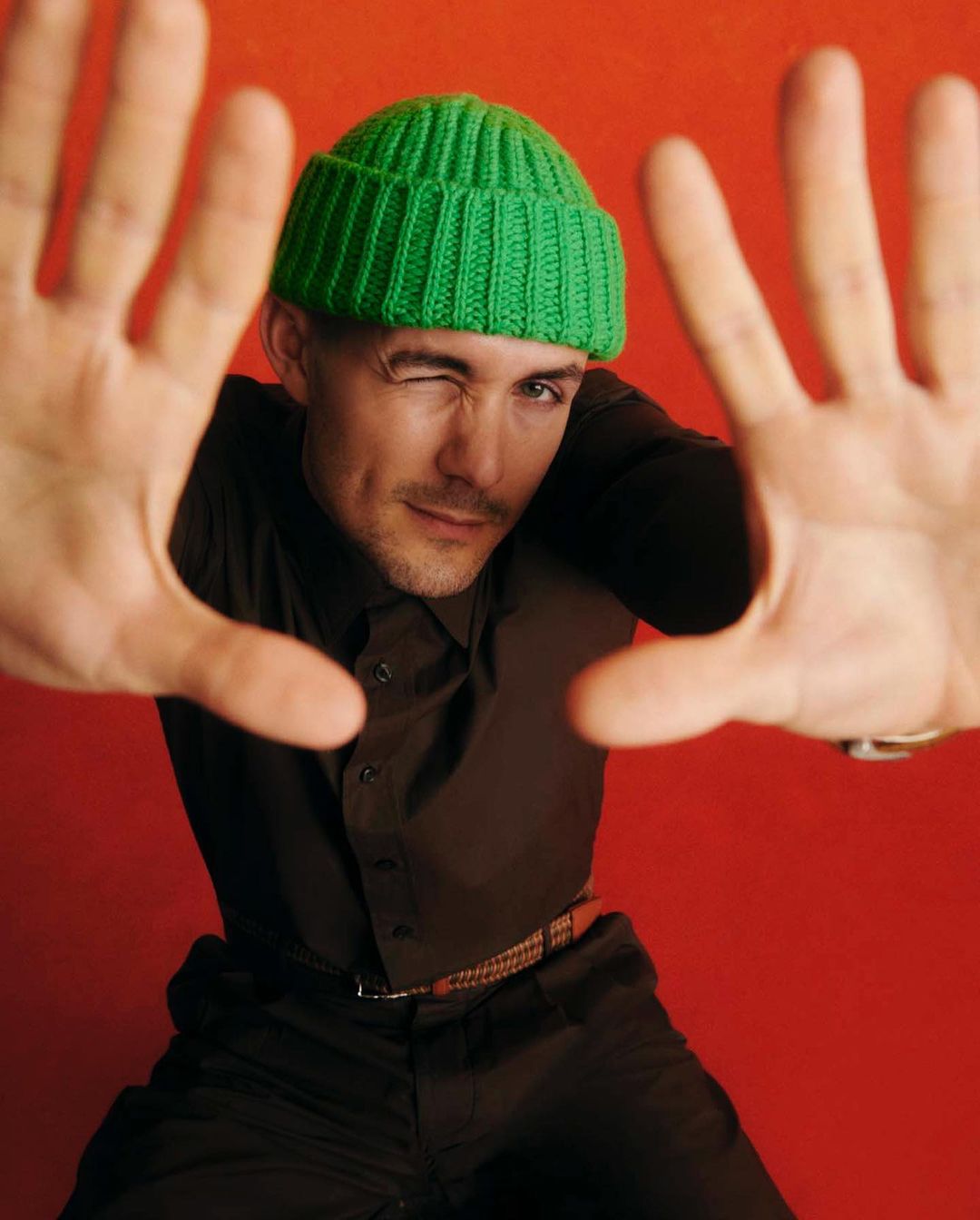 Man wearing a green beanie and dark shirt holding both hands up toward the camera against a textured red background
