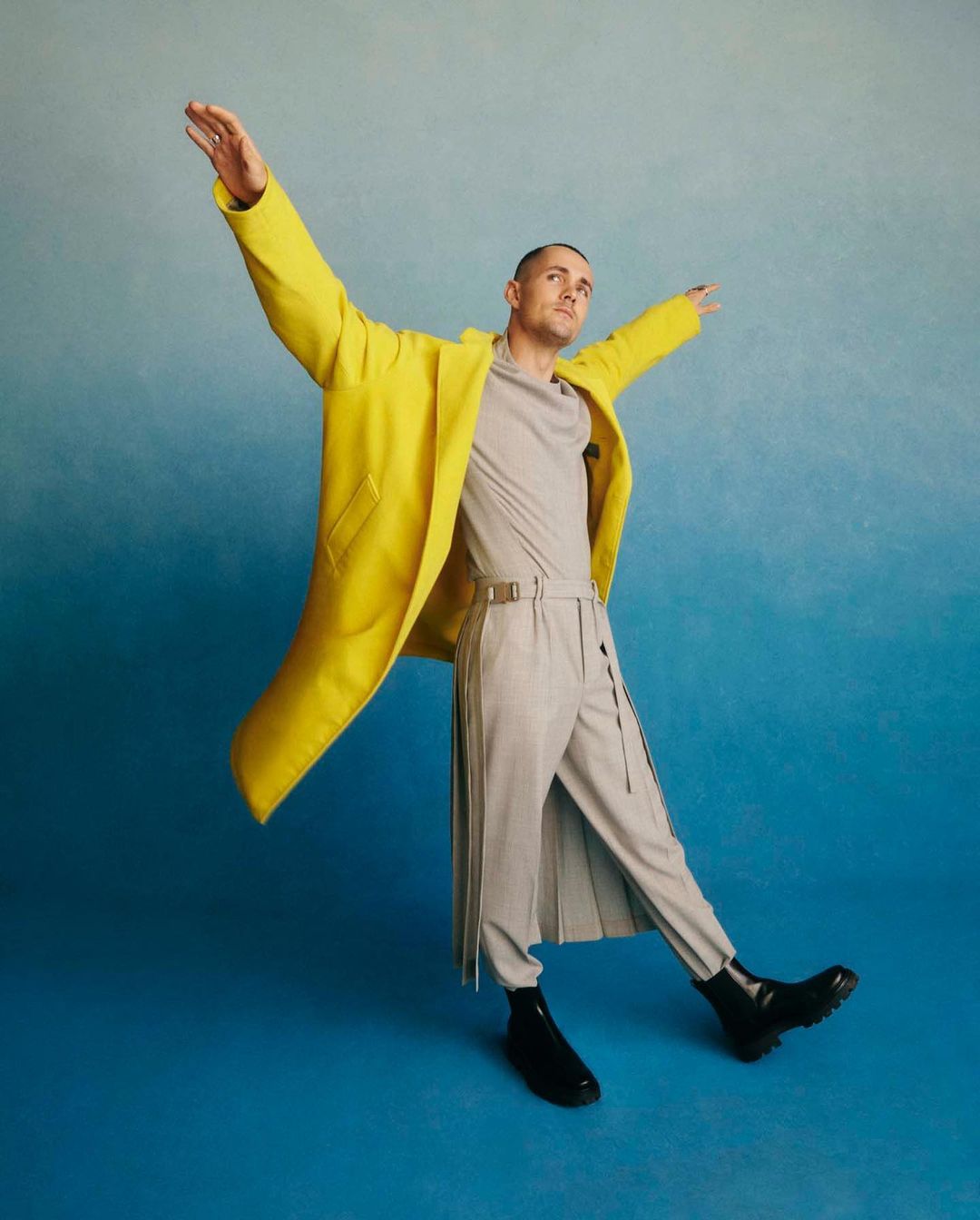 A man in a bright yellow coat poses with arms outstretched against a graduated blue background, creating a dynamic and stylish fashion image.