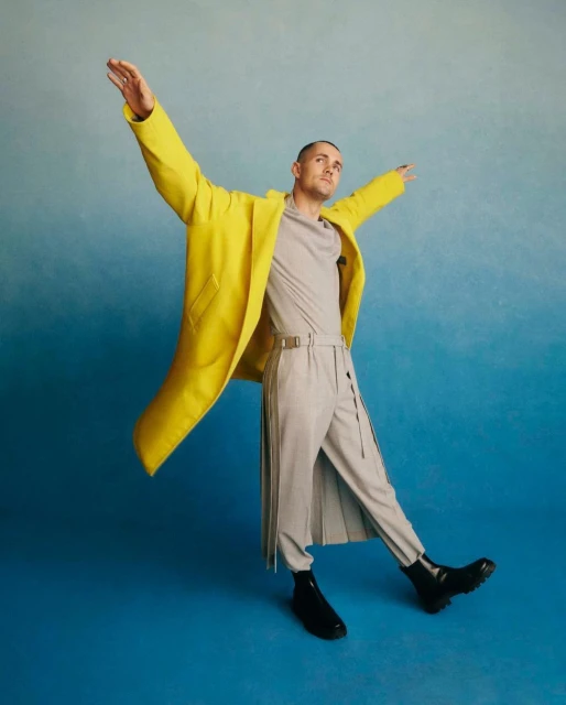 A man in a bright yellow coat poses with arms outstretched against a graduated blue background, creating a dynamic and stylish fashion image.