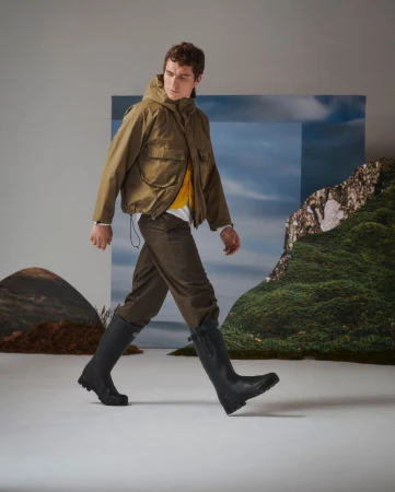 Man walking in a studio wearing a hooded olive jacket, brown trousers, and tall black rubber boots.