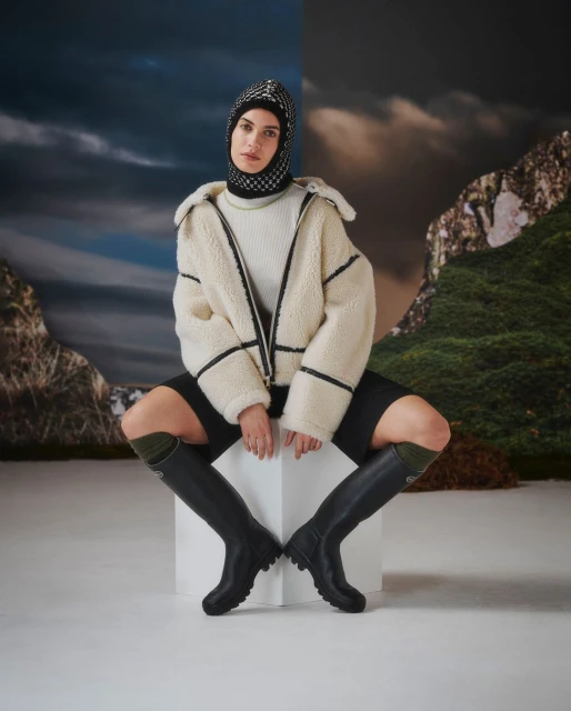 Fashionable woman in stylish jacket, balaclava, and knee-high boots, seated on a white cube against a scenic mountain backdrop.