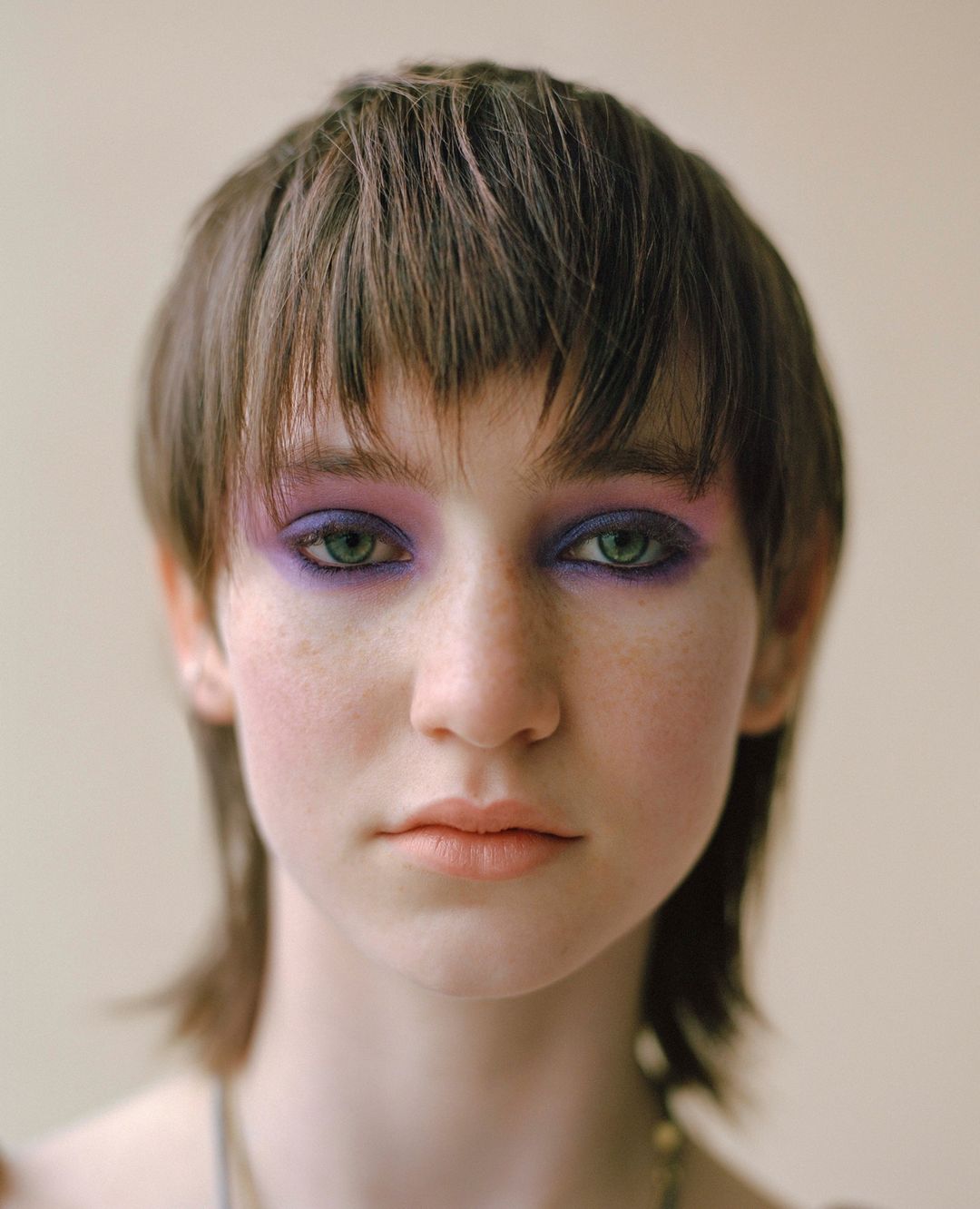 Close-up studio portrait of a young adult female with a dark brown mullet and purple eyeshadow, set against a neutral beige background.