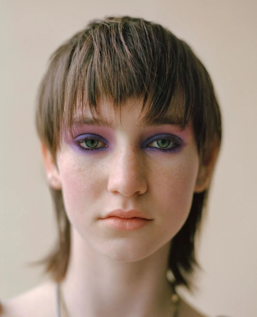 Close-up studio portrait of a young adult female with a dark brown mullet and purple eyeshadow, set against a neutral beige background.